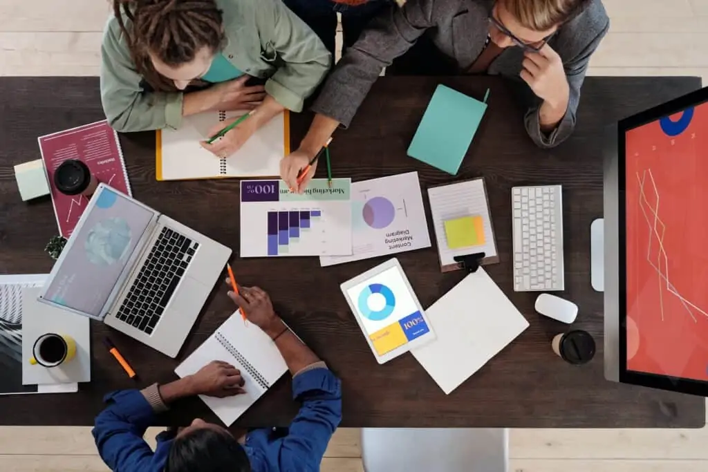 Three people at a meeting table with a laptop, documents, and graphs