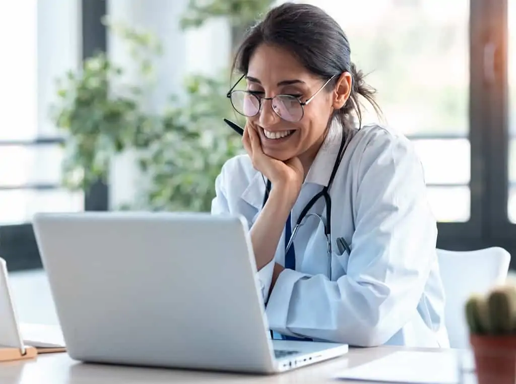 A female medical professional wearing glasses smiling at her laptop