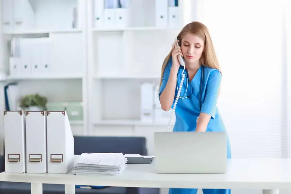 A medical professional in blue scrubs answering the phone at her desk